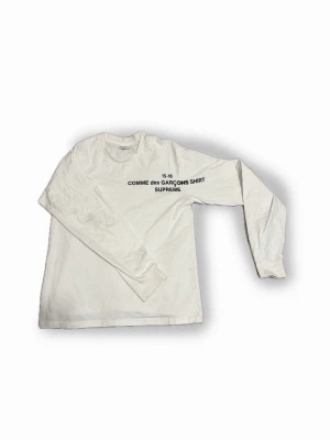 Vit långärmad Supreme x Comme des Garçons - Vit långärmad t shirt med tryck 'COMME des GARÇONS SHIRT SUPREME' på bröstet. Klassisk rund hals och rak passform. Sitter som en S