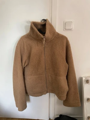 Beige teddyjacka med dragkedja - Mysig brun/beige teddyjacka i fluffigt material med hög krage och hel dragkedja framtill. Jackan har två stora fickor och en avslappnad passform.