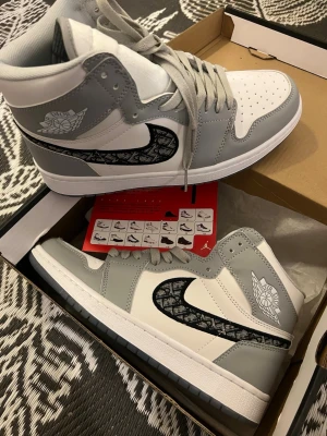 Gråvita Air Jordan 1 High sneakers - Säljer ett par gråvita Air Jordan 1 High sneakers med svart Swoosh-detalj i mönstrad design. Skorna har högt skaft, vita paneler och gråa partier i läder. Klassisk Air Jordan-logga på sidan och grå snörning. Perfekta för dig som gillar streetwear och snygga detaljer.