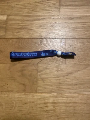 Blått Sverigedemokraterna armband - Säljer ett blått tygarmband med texten 'Sverigedemokraterna' i ljusblått, dekorerat med små blommor och bin. Armbandet har en vit pärla som lås och är gjort av tygband.