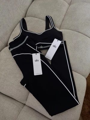 Alo Yoga Set - Snyggt svart träningsset från Alo med sport-bh och tights. Setet har vita kontrastlinjer och logga på både bh och byxa. Perfekt för gymmet eller yoga. Materialet är stretchigt och följsamt för maximal komfort.