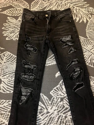 Svarta slitna jeans med hål - Säljer ett par svarta jeans med flera stora slitningar och hål framtill på båda benen. Jeansen har fem fickor, knappgylf och en smal passform. Perfekta för en edgy och trendig look.