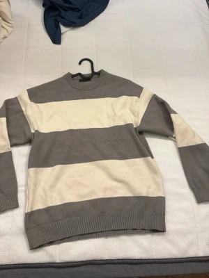 Randig beige och grå stickad tröja - Stickad tröja med breda horisontella ränder i beige och grått. Tröjan har rund halsringning, långa ärmar och ribbade muddar vid ärmslut och nederkant. Perfekt för dig som gillar en avslappnad och stilren look.