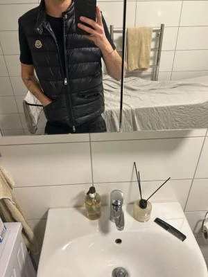 Svart dunväst från Moncler - Snygg svart dunväst från Moncler med glansig finish och klassisk logotyp på bröstet. Västen har ståkrage, dragkedja framtill och två fickor med dragkedja. Perfekt för lager-på-lager och håller dig varm under kyliga dagar. Köpt från Fashionels, storlek 2 i moncler storlek!