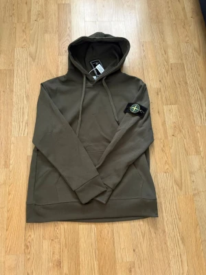 Olivgrön hoodie från Stone Island - Säljer en olivgrön hoodie från Stone Island med klassisk patch på vänster ärm. Hoodien har huva med dragsko och en stor magficka framtill. Tillverkad i mjukt bomullsmaterial och har ribbade muddar vid ärmslut och nederkant. Perfekt för en avslappnad streetwear-look.storlek S men passar typ 168cm