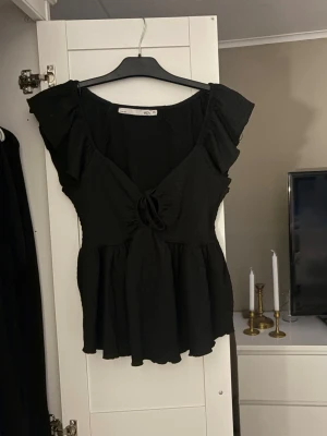 Svart blus med volang och knyt - Snygg svart blus med volangdetaljer på axlarna och v-ringning med knyt framtill. Blusen har en luftig passform och är tillverkad i ett mjukt tyg som ger ett fint fall. Perfekt att styla med jeans eller kjol för en trendig look.
