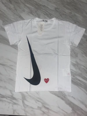 CDG T-shirt - Snygg vit t-shirt från Comme des Garçons Play med stor svart Nike-liknande swoosh och det klassiska röda hjärtat med ögon. Framsidan har även PLAY-logga tryckt i svart. Rund hals och korta ärmar, tillverkad i mjuk bomull. Digital KVITTO från Sellpy finns KOLLA MÅTTEN NOGGRANT 