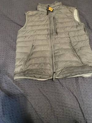 Grå dunväst från Parajumpers - Snygg grå dunväst från Parajumpers i storlek S. Västen är superlätt, har dragkedja framtill, två sidofickor med dragkedja och en bröstficka. Perfekt för lager-på-lager och har klassisk quiltad design med hög krage och gul detalj vid halsen. Kan också byta mot något 