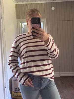 Randig långärmad tröja i vitt och vinrött - Säljer en oversized långärmad tröja med breda vinröda och vita ränder. Tröjan har rund halsringning och avslappnad passform, perfekt för en chill och enkel stil. Materialet ser ut att vara mjuk bomull.