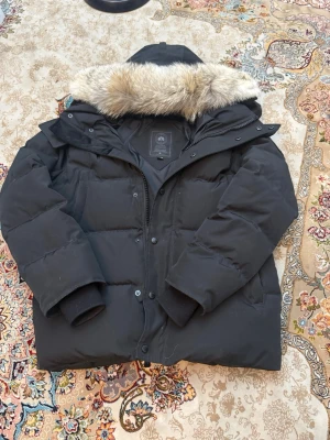 Svart Canada Goose jacka - Säljer en svart dunjacka från Canada Goose med stor huva och avtagbar pälskant. Jackan har klassisk patch på ärmen, dragkedja och knappar framtill samt breda muddar vid ärmsluten. Perfekt för kalla vinterdagar och riktigt varm.