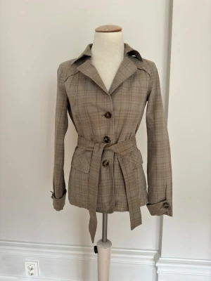 Beige rutig jacka med bälte - Snygg beige jacka med rutigt mönster, knappar framtill och matchande bälte i midjan. Jackan har klassisk krage och långa ärmar, perfekt för dig som vill ha en tidlös och clean look till vardags. Materialet känns mjukt och lätt.