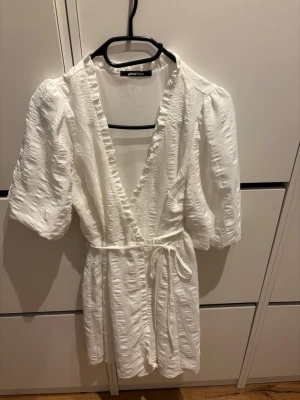 Vit omlottklänning från Gina Tricot - Supersöt vit omlottklänning från Gina Tricot med puffiga korta ärmar, volangkant längs v-ringningen och knytband i midjan. Perfekt för sommarens alla tillfällen och ger en fräsch och trendig look. Passar perfekt till även skolavslutning eller konfirmation. Använd endast en gång på min konfirmation. 
