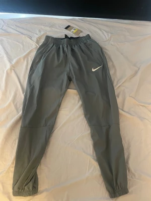 Gråa träningsbyxor från Nike - Snygga gråa mjukisbyxor från Nike med elastisk midja och muddar vid bensluten. Byxorna har en enkel design med vit Nike-logga på ena benet och är tillverkade i ett lätt och bekvämt material. Perfekta för chill eller träning.