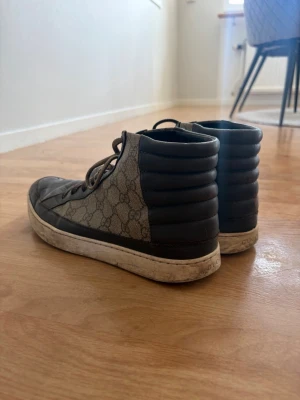 Bruna high top sneakers - Snygga high top sneakers i brunt läder och canvas med det klassiska GG-mönstret. Skorna har vadderad häl, bruna snören och vit sula. Perfekta för dig som vill ha en lyxig och trendig look.