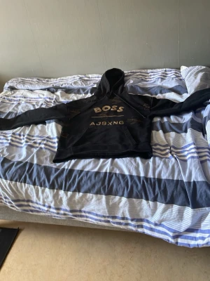 Svart hoodie med guldtext BOSS - Snygg svart hoodie med huva och dragsnören. Stor BOSS-logga i guld på bröstet och guldfärgade signaturer tryckta runtom. Hoodien har en klassisk passform och känguruficka framtill. Perfekt för dig som vill sticka ut med stilren streetwear.