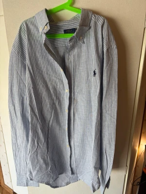 Ralph Lauren skjorta - Klassisk långärmad skjorta med blåvita smala ränder och button-down krage. Skjortan har broderad logga på bröstet och vita knappar framtill. Tillverkad i ett lätt bomullstyg som passar perfekt till en avslappnad men ändå stilren look. Skjortan är helt oanvänd och bara testad men storleken passade ej.
