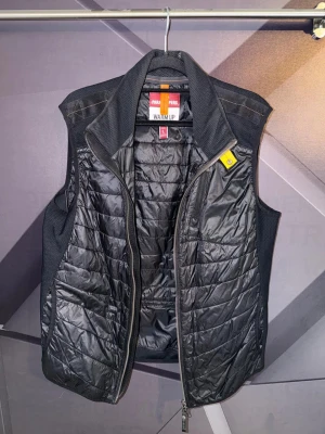 Parajumpers väst - Snygg svart dunväst från Parajumpers med quiltad design och hög krage. Västen har dragkedja framtill och detaljer i gult vid halsen. Perfekt för lager-på-lager och håller dig varm under kyliga dagar.