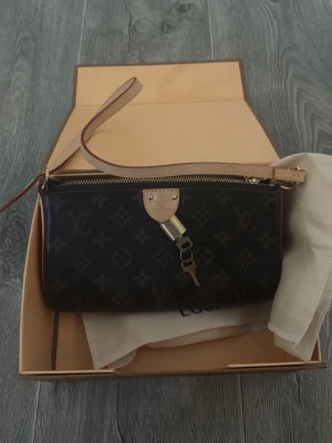 Louis Vuitton monogram clutch - Säljer en snygg mörkbrun clutch från Louis Vuitton med klassiskt monogrammönster och guldfärgade detaljer. Väskan har en dragkedja, beige läderrem och dekorativt hänglås. Perfekt för att bära det viktigaste och ge en lyxig touch till din outfit. Köpt 2022 den  är använd men fortfarande i väldigt bra skick. Har dessvärre inget kvitto då detta var en present . 