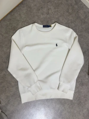 Vit sweatshirt från Polo Ralph Lauren - Klassisk vit sweatshirt från Polo Ralph Lauren med rund halsringning och broderad marinblå logga på bröstet. Tröjan har ribbade muddar vid ärmslut och nederkant samt en diskret V-detalj vid halsen. Perfekt för en clean och stilren look.