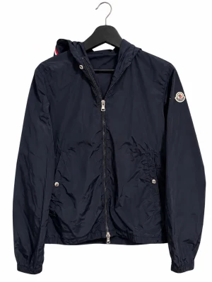 Moncler Grimpeurs windbreaker Navy – Size 1 - Moncler Grimpeurs windbreaker i mörkblå/navy, storlek 1. Snöret har tyvärr tappats bort. Lätt vattenavvisande nylonjacka med huva, tvåvägsdragkedja, tryckknappsfickor, elastiska avslut och Monclers klassiska tricolor-detalj samt ärmlogo. Stilren och perfekt vår/höst. Fint begagnat skick enligt bilder. Originaletiketter visas. Mått skickas gärna. Bud välkomnas & snabb frakt 📦