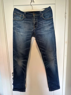 Blå slim fit jeans från Nudie Jeans - Snygga blå slim fit jeans från Nudie Jeans med klassisk femficksmodell och slitningar på framsidan. Jeansen har en mellanblå tvätt med ljusare partier och kontrastsömmar. Bakfickorna har Nudie Jeans karakteristiska broderi. Perfekta för en avslappnad stil.