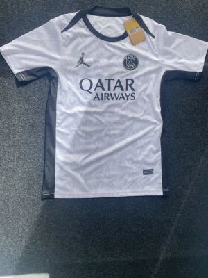 PSG Träningströja 26/27 - Snygg PSG matchtröja i vitt med svarta detaljer och logga från Jordan. Tröjan har korta ärmar, svart krage och Qatar Airways tryckt på bröstet. Tillverkad i lätt och ventilerande material, perfekt för fotboll eller träning.