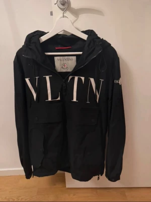 Svart vindjacka Valentino x Moncler - Snygg svart vindjacka med stor vit VLTN-logga framtill. Jackan har huva med dragsko, två bröstfickor med lock och dragkedja framtill. Märkt med både Valentino och Moncler på insidan. Perfekt för dig som gillar exklusiva samarbeten. Jackan har ett hål i armhålan (se sista bilden). Lagning kostar ca 200–300 kr. Annars är jackan i väldigt bra skick.