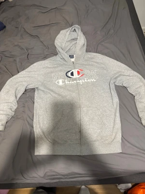 Grå zip hoodie från Champion XL - Snygg grå zip hoodie från Champion i storlek XL. Hoodien har dragkedja, två fickor fram och tryck med texten 'New York Champion' samt Champion-loggan på bröstet. Tillverkad i mjukt bomullsmaterial och har en klassisk huva.