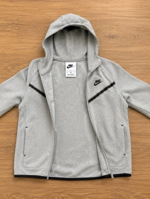 Grey Nike hoodie with zipper  - Snygg grå hoodie från Nike med hel dragkedja och svarta detaljer över bröstet. Hoodien har huva, två fickor framtill och Nike-logga på vänster sida. Perfekt för en sportig och avslappnad stil.