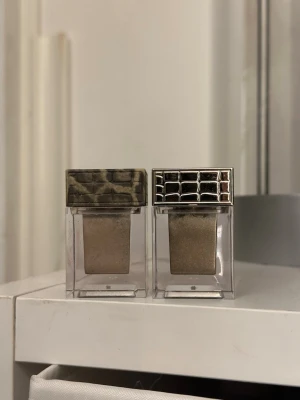 Två ögonskuggor från Make Up Store - Swatchade. Säljer två ögonskuggor från Make Up Store i fyrkantiga genomskinliga burkar. Ena har lock med leopardmönster och den andra har ett silvrigt lock med rutmönster. Båda skuggorna är i en skimrande beige/guldig nyans.