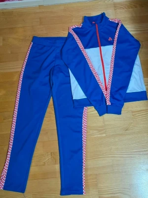 Blått träningsset för barn  - Sportigt träningsset i blått och vitt med röda och vita rutiga detaljer längs ärmar och byxben. Jackan har hög krage, hel dragkedja i rött och logga på bröstet. Tillverkat i ett mjukt syntetmaterial som passar perfekt för träning eller chill.