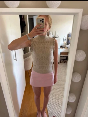 Beige stickad ärmlös topp - Säljer en beige stickad topp med hög krage och ärmlös design. Toppen har ett fint hålmönster och bred ribbad kant nedtill. Perfekt för dig som gillar en stilren och trendig look.