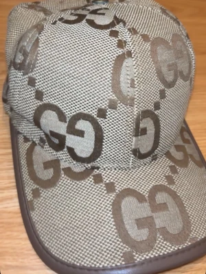Beige Gucci keps - Snygg beige keps med brunt GG-mönster över hela ytan och bruna detaljer längs skärmen. Kepsen har en klassisk form och är tillverkad i ett tåligt tygmaterial. Perfekt för dig som gillar stilrena accessoarer med tydlig logga.