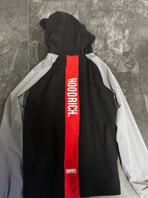 Svart och grå vindjacka från Hoodrich - Snygg vindjacka från Hoodrich i svart och grått med röda detaljer. Jackan har huva, dragkedja framtill och en röd ficka på magen. Stor Hoodrich-logga på ryggen och ärmen. Perfekt för streetwear-stil.