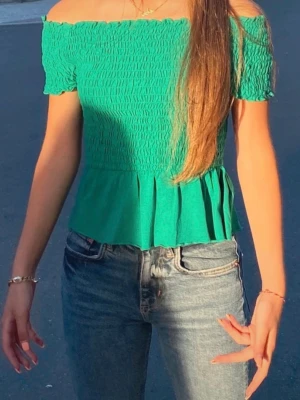 Grön offshoulder topp med volang - Snygg grön offshoulder topp med smockad överdel och volang nedtill. Toppen är kortärmad och har en figurnära passform upptill som släpper ut i en volang. Perfekt till jeans eller kjol för en trendig look.