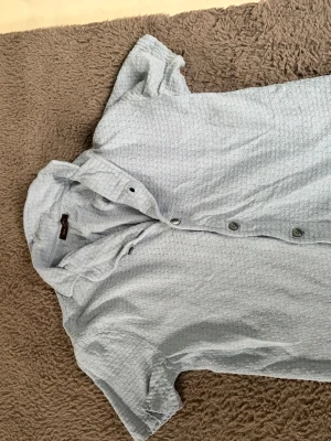 Ljusblå kortärmad henley skjorta - Säljer en ljusblå henley skjorta med korta ärmar och knappar framtill. Skjortan har ett diskret strukturerat mönster i tyget och är tillverkad i ett mjukt material som känns skönt mot huden. Perfekt för dig som gillar en avslappnad men ändå stilren look.