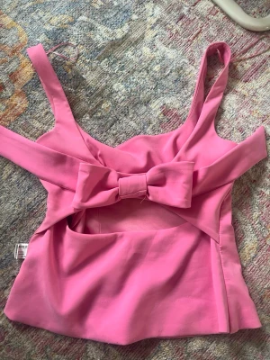 Rosa linnetopp med rosett och cut out - Supersöt rosa linnetopp med breda axelband, stor rosettdetalj baktill och cut out under bysten. Toppen har en figurnära passform och är perfekt för dig som vill sticka ut lite extra. Materialet känns mjukt och följsamt.