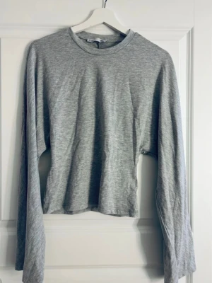 Grå långärmad figursydd tröja - Enkel och stilren grå långärmad figursydd tröja i mjukt bomullsmaterial och utsvängda ärmar. Tröjan har rund halsringning och rak passform, perfekt att matcha med jeans eller kjol. Passar dig som gillar minimalistisk och avslappnad stil.