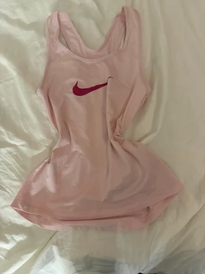Ljusrosa Nike Dri-FIT linne - Säljer ett ljusrosa träningslinne från Nike med Dri-FIT material som andas. Linnet har en racerback-design och en mörkrosa Nike-logga på bröstet. Perfekt för gymmet eller löpning när du vill hålla dig sval och bekväm.
