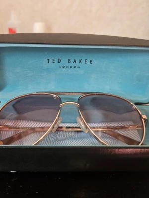 Ted Baker aviator solglasögon guld - Snygga aviator solglasögon från Ted Baker med guldfärgad metallbåge och mörktonade glas. Klassisk och stilren design som passar perfekt till sommaren. Kommer med originalfodral.