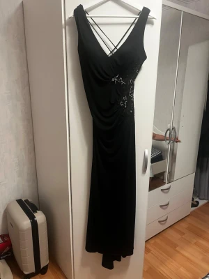 Svart långklänning med spetsdetaljer - Elegant svart långklänning med v-ringning och smala axelband som korsas i ryggen. Klänningen har fina spetsdetaljer med pärlor på sidan och lätt drapering framtill. Perfekt för dig som vill sticka ut med en stilren look. As snygg klänning för soeciella tillfällen. Använd en gång och köpt ifrån USA. 
