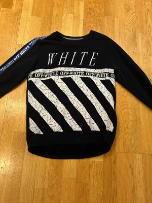 Svart Off-White hoodie med tryck - Säljer en svart hoodie från Off-White med stora vita diagonalränder och texten 'WHITE' på bröstet. Har även Off-White-logga på bröstet och längs ena ärmen. Hoodien har en modern, oversized passform och är tillverkad i mjukt bomullsmaterial.