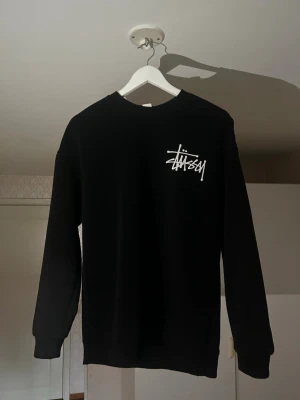Svart sweatshirt från Stüssy - Svart sweatshirt från Stüssy med klassisk vit logga tryckt på bröstet. Tröjan har rund halsringning, långa ärmar och ribbade muddar vid ärmslut och nederkant. Perfekt för en streetwear-look och enkel att matcha med jeans eller byxor.