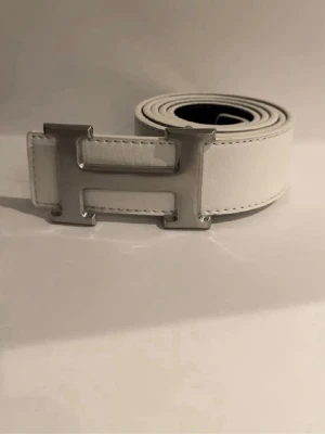 H bälte 110cm Vitt skärp med stor silverspänne - Snyggt vitt skärp i skinn med ett stort, blankt silverspänne i form av ett H. Perfekt accessoar för att lyfta din outfit och ge en clean look. Skärpet har stilrena sömmar och känns riktigt modernt.