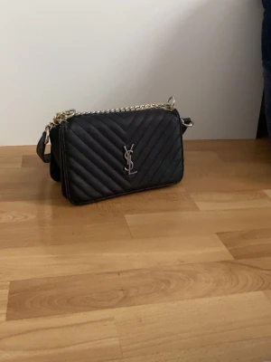 Svart YSL quiltad axelremsväska - Snygg svart quiltad handväska från YSL med silverfärgad kedja och logga framtill. Väskan har en stilren, rektangulär form och är tillverkad i läder. Perfekt för dig som vill ha en trendig och lyxig accessoar.