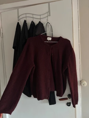 Vinröd stickad kofta med knappar - Mysig vinröd stickad kofta med rund halsringning och knäppning framtill. Koftan har långa ärmar och en rak passform, perfekt att slänga över en t-shirt. 
