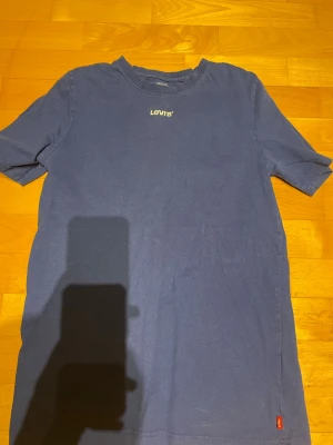 Blå t-shirt från Levi's - En klassisk blå t-shirt från Levi's med liten vit logga på bröstet och röd etikett vid nederkanten. T-shirten har rund hals och korta ärmar, perfekt för en avslappnad stil.