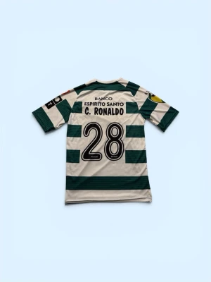 Grönvit fotbollströja Sporting CP - Säljer en klassisk grönvit randig fotbollströja från Sporting CP med nummer 28 och namnet C. Ronaldo på ryggen. Tröjan har korta ärmar, klubbmärke på bröstet och flera sponsorloggor. Perfekt för dig som älskar fotboll och vill sticka ut på planen.