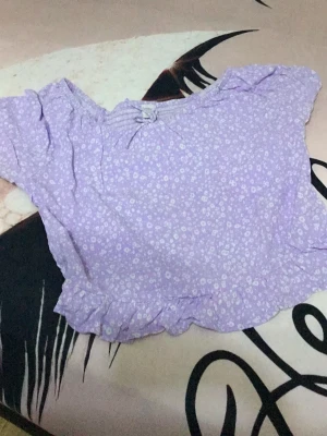 Ljuslila blommig croppad blus - Söt ljuslila blus med små vita blommor och volangkant nertill. Modellen är croppad och har korta ärmar. Perfekt för dig som gillar romantisk och trendig stil.