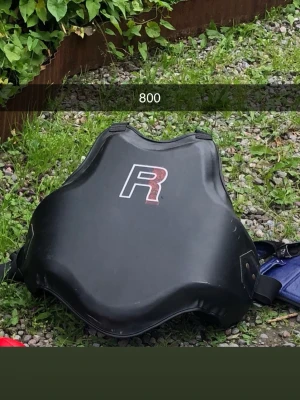 Svart boxningsväst med R-logga - Säljer en svart boxningsväst med stor vit och röd R-logga på bröstet. Västen har justerbara remmar på sidorna och är tillverkad i slitstarkt syntetmaterial, perfekt för träning och sparring inom kampsport.
